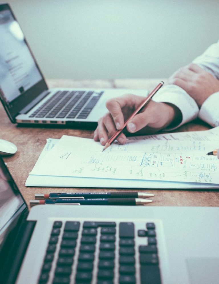 Händed mit Stift über Notizen, Laptops und Schreibmaterialien auf einem Tisch.
